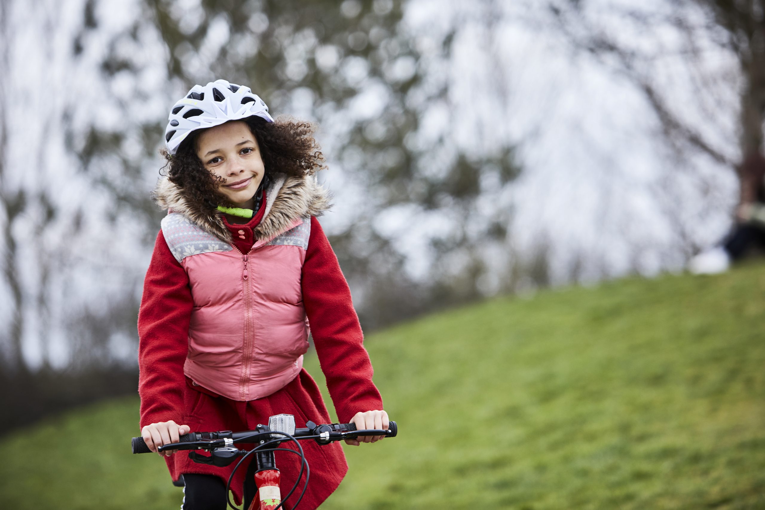 A young girl on her cycle in a park. She has a pink body warmer on over a red coat and a big grin