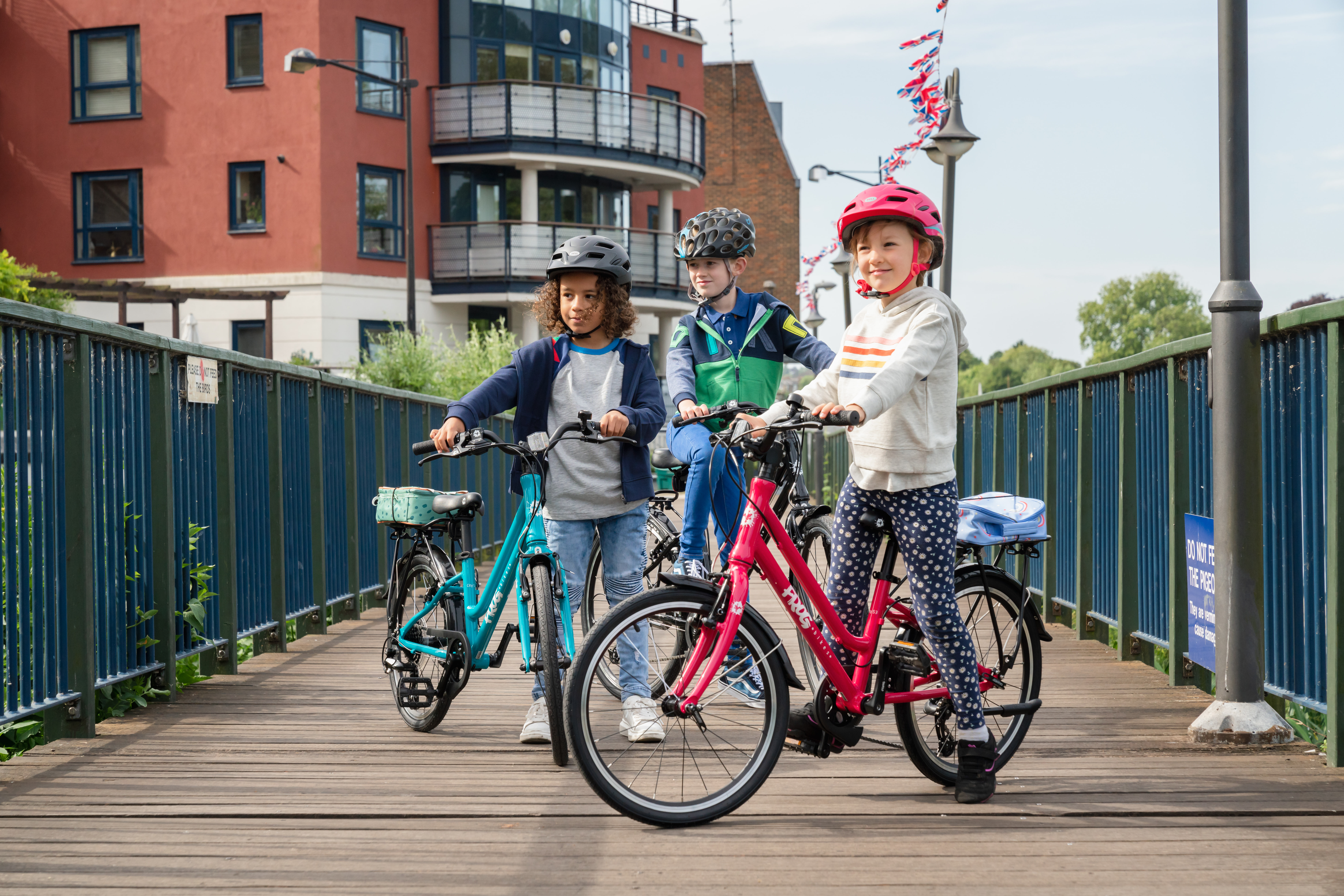 Three children on their bicycles on a bridge. They are wearing helmets and smiling