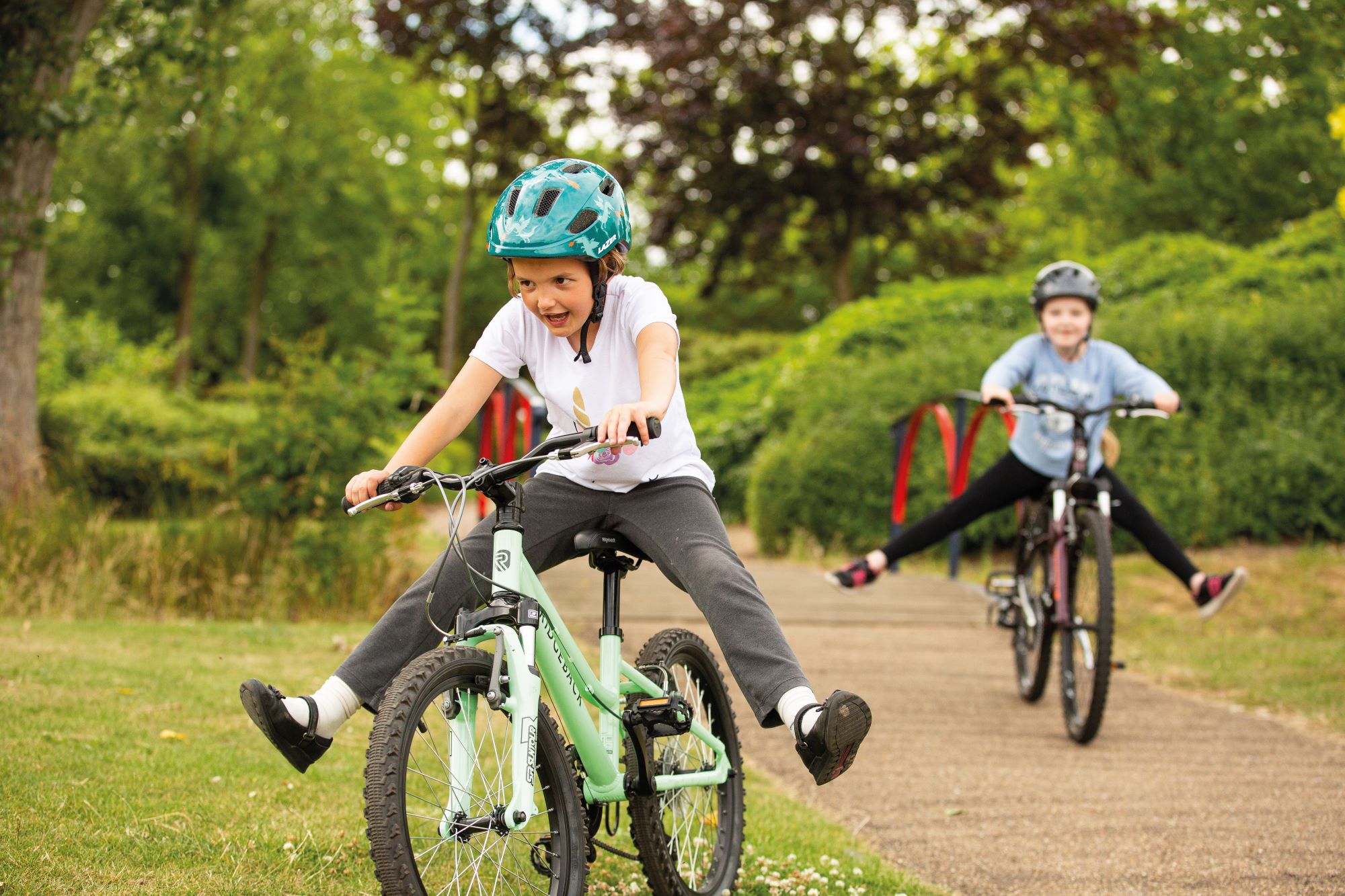 Two children cycling in a park. They are laughing and have their legs sticking out as they freewheel