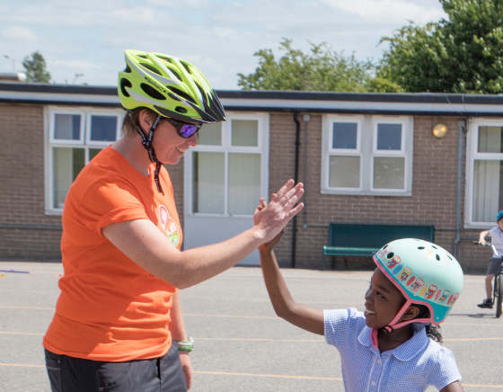 A Bikeability instructor wearing an oragne t shirt high fives a young girl on a bike wearing a checked school dress and helmet.