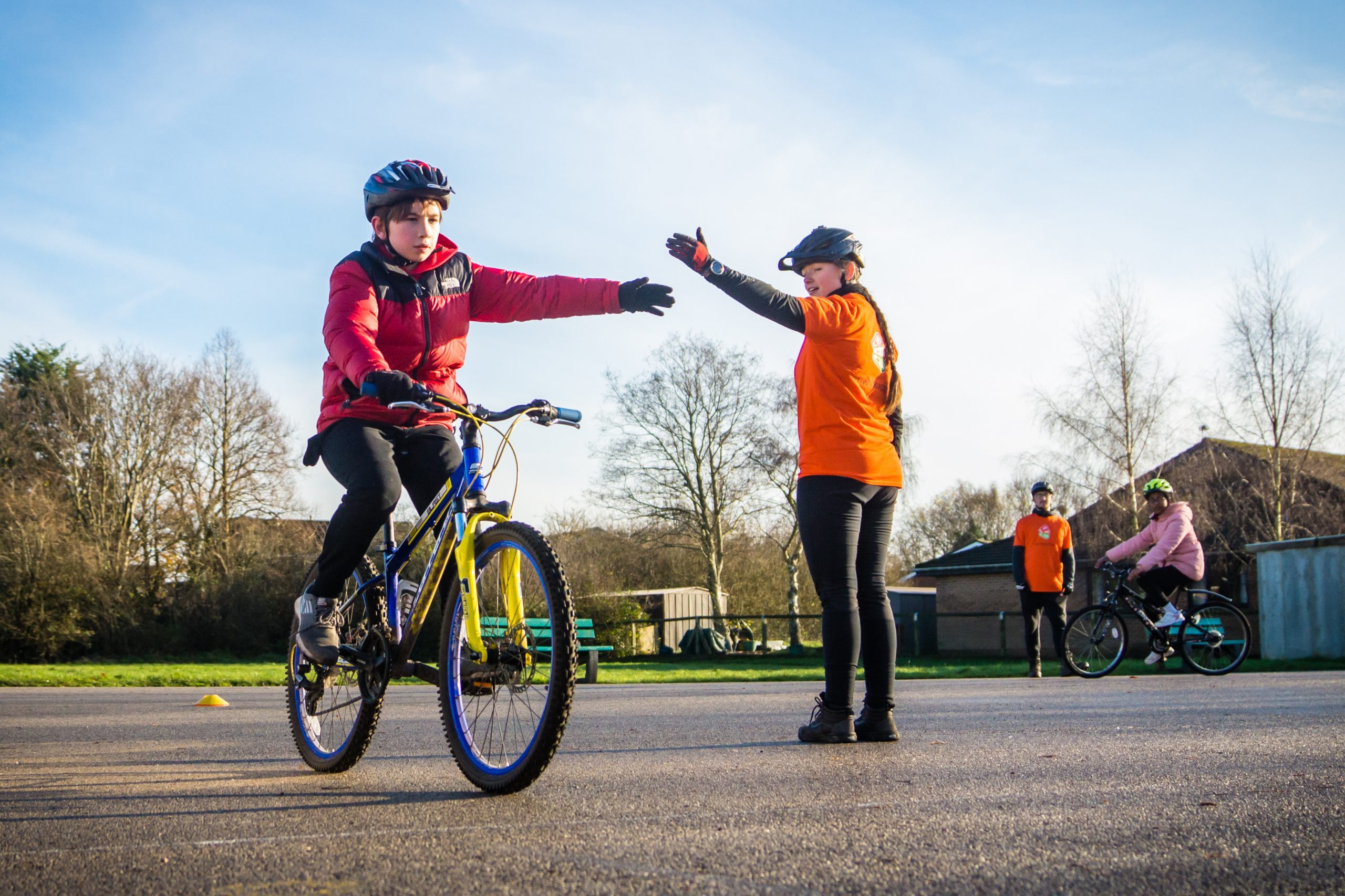 A boy on a bike reaches out to signal with his left arm. A female instructor reaches out her arm to show him what to do.