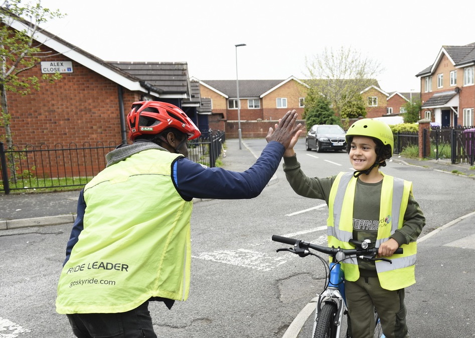 A male Bikeability instructor wearing a high vis jacket high fives a young boy stood next to a cycle.