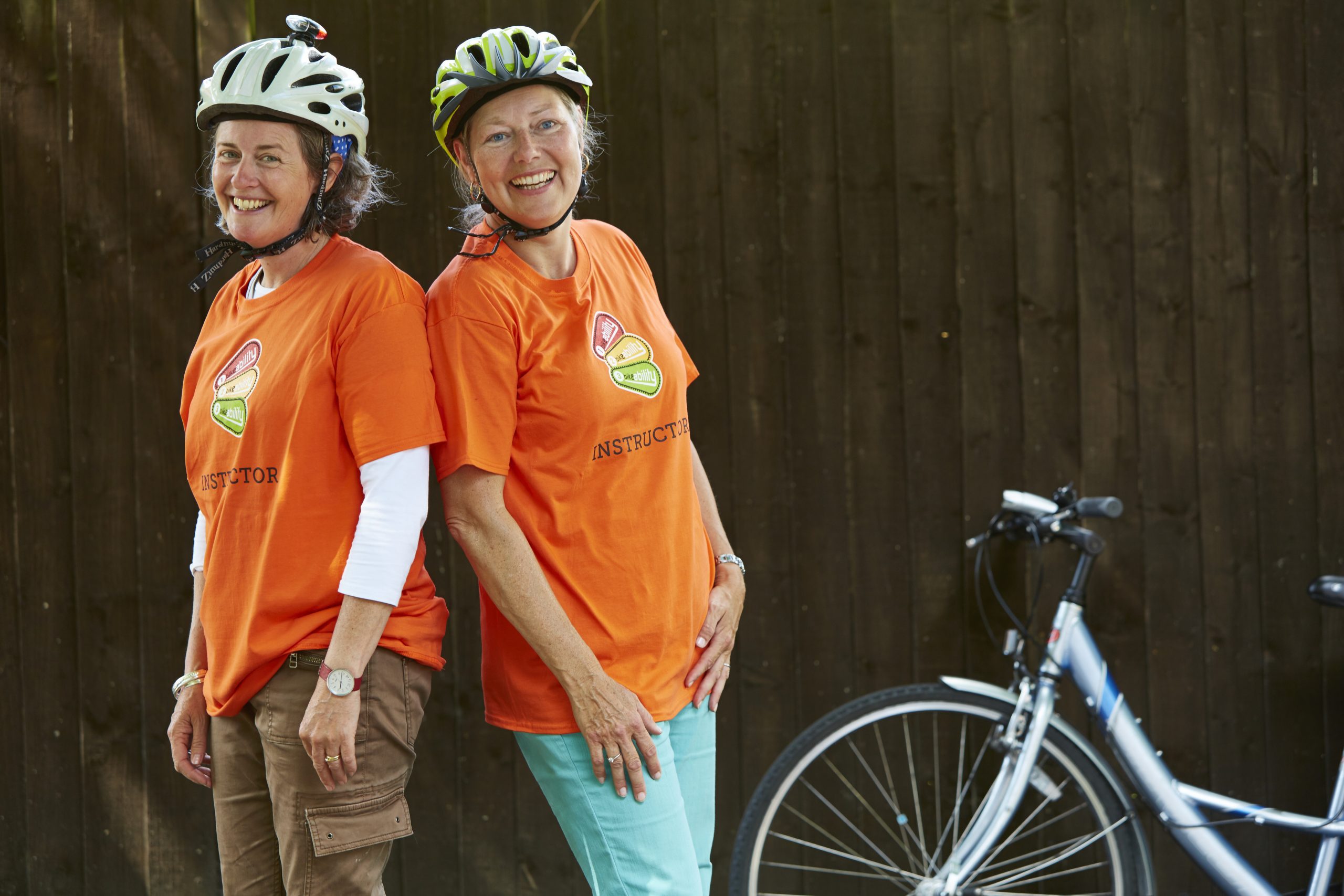 Two female Bikeability instructors wearing orange t shirts stanfd back to back and smiling next to a cycle.