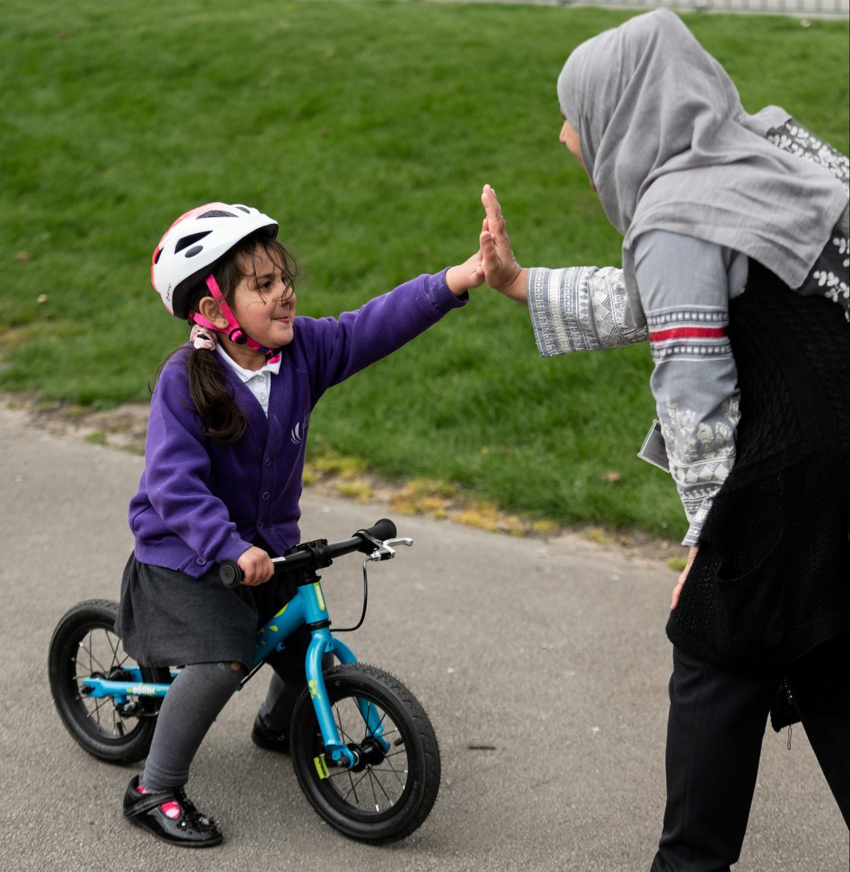 A young girl riding a balance bike high fives a woman on a playground.