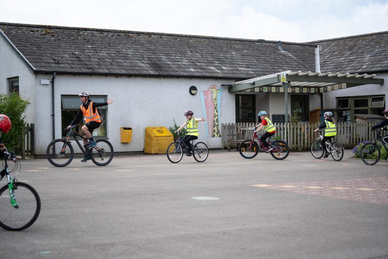 A Bikeability instructor leads children around the playground on cycles during Bikeability 