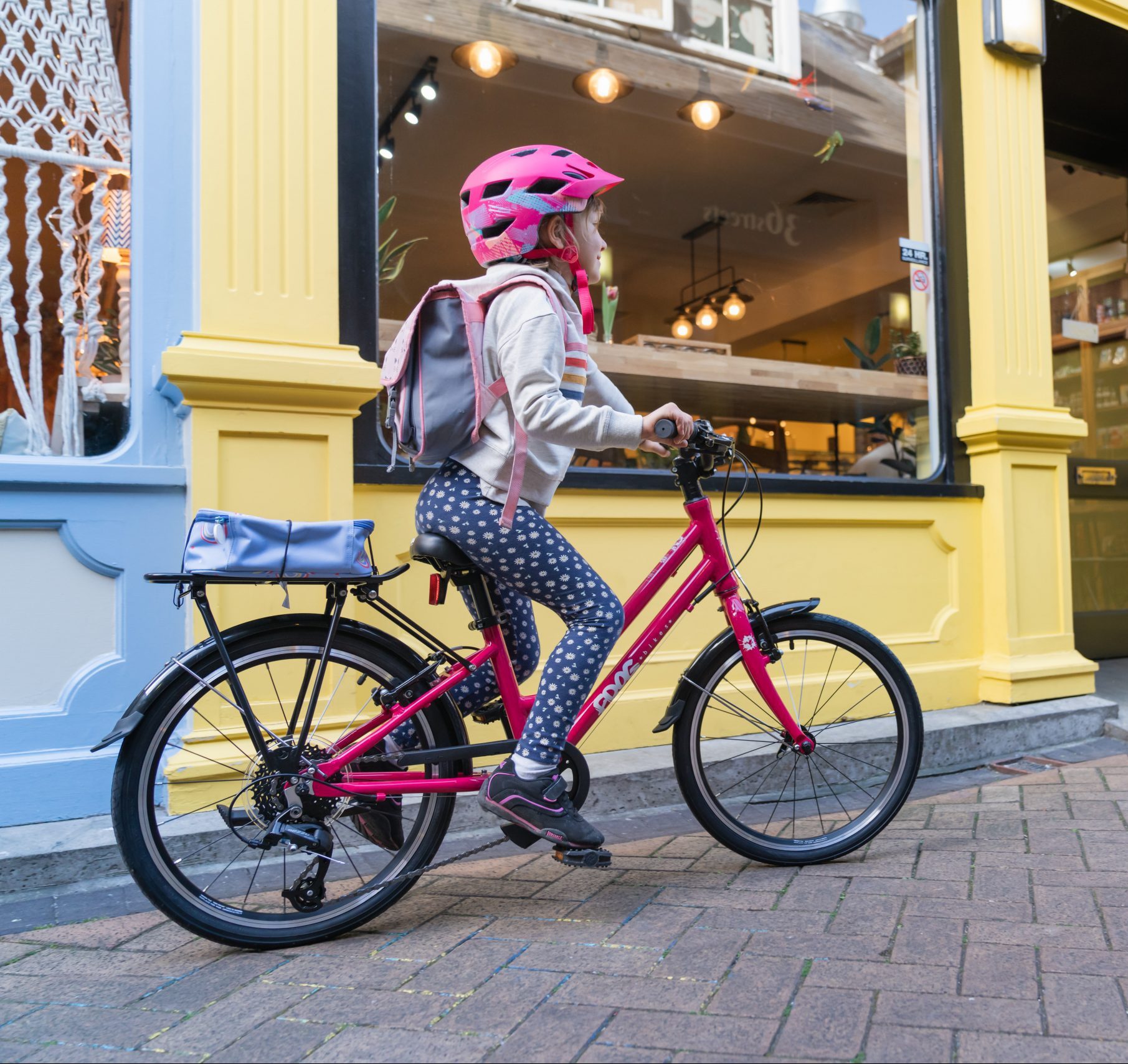 A child wearing a backpack and a pink helmet cycling on a red cycle on a high street