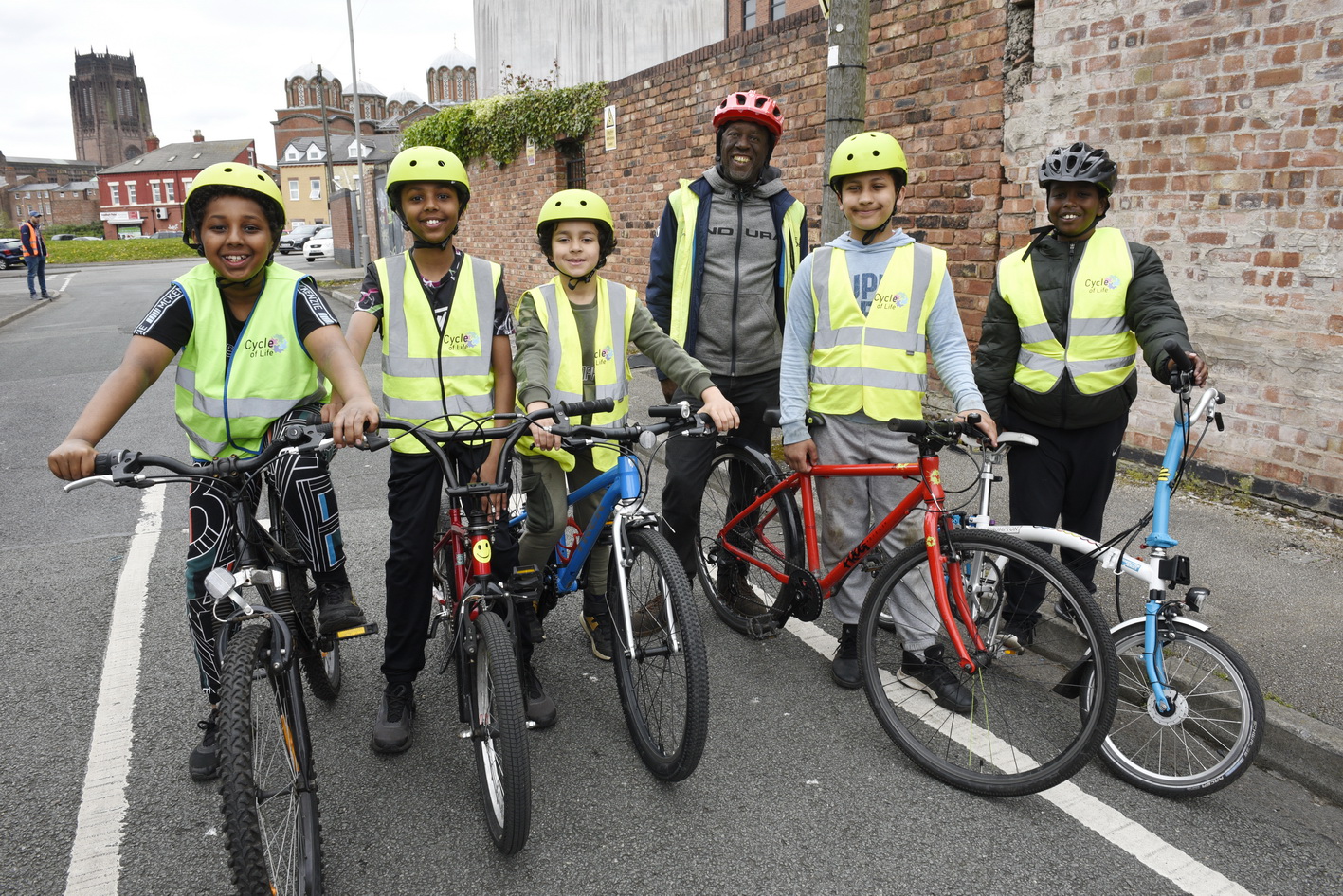 A group of young cyclists are standing on or with their bicycles. They are wearing Cycle of Life high vis vests, and smiling. Behind them is a Bikeability instructor.