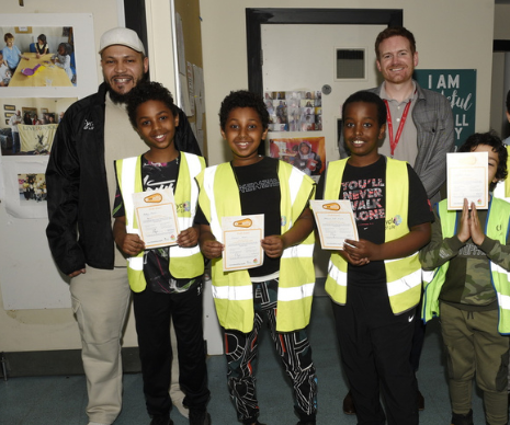 Three young boys smiling, wearing high vis vest and holiding Bikeability certificates stand in front of two men.
