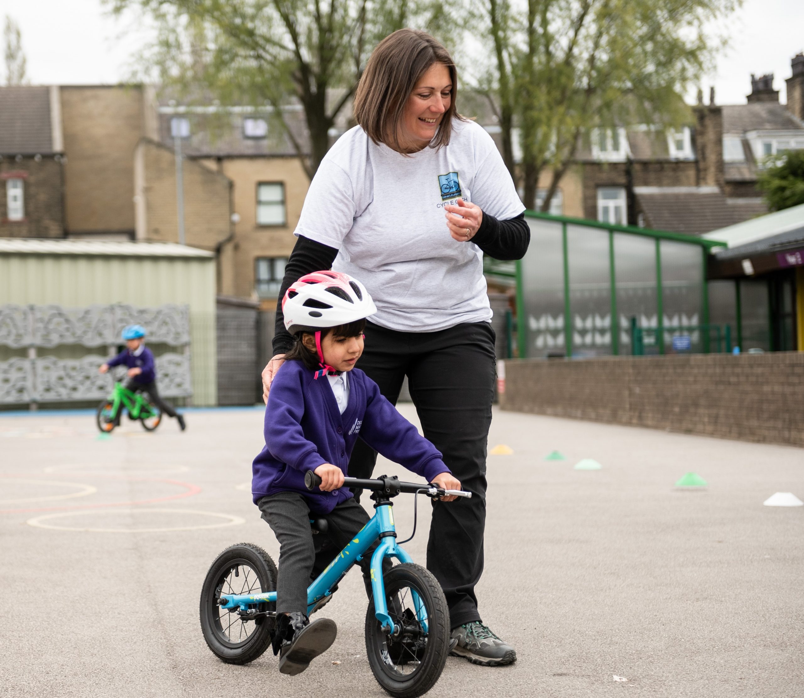 A young girl in a purple school cardigan is on a balance bike whilst a woman in a white t shirt guides her. They are both on a playground.