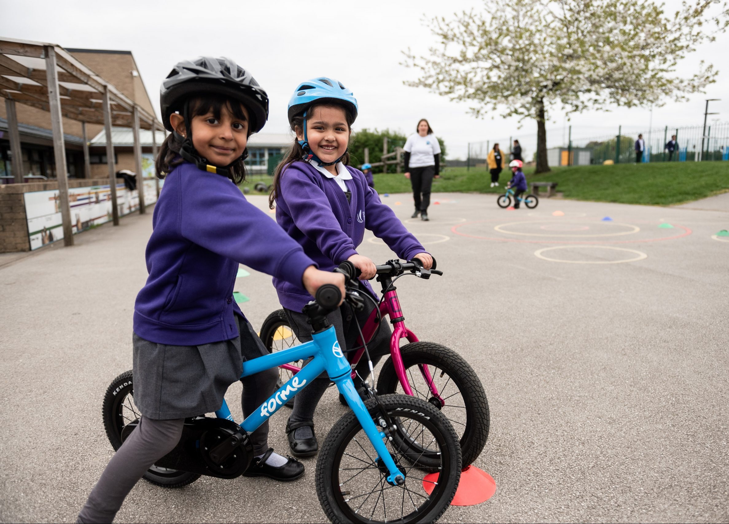 Two young girls wearing purple school cardigans and helmets smile at the camera as they straddle balance bikes.