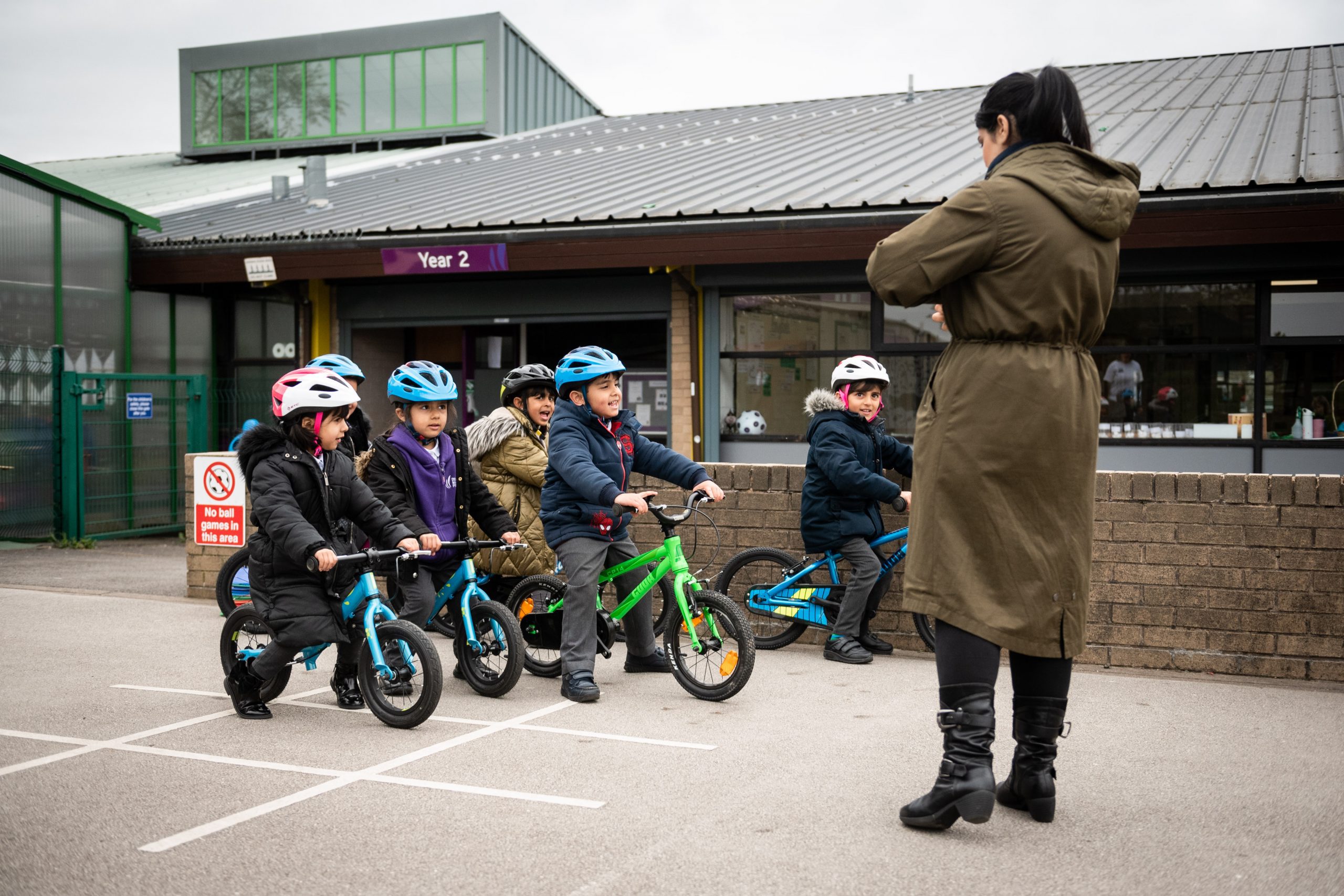 A group of five small children stand on balance bikes in a playground. A woman wearing a green parka stands with her back to the camera, teaching them.