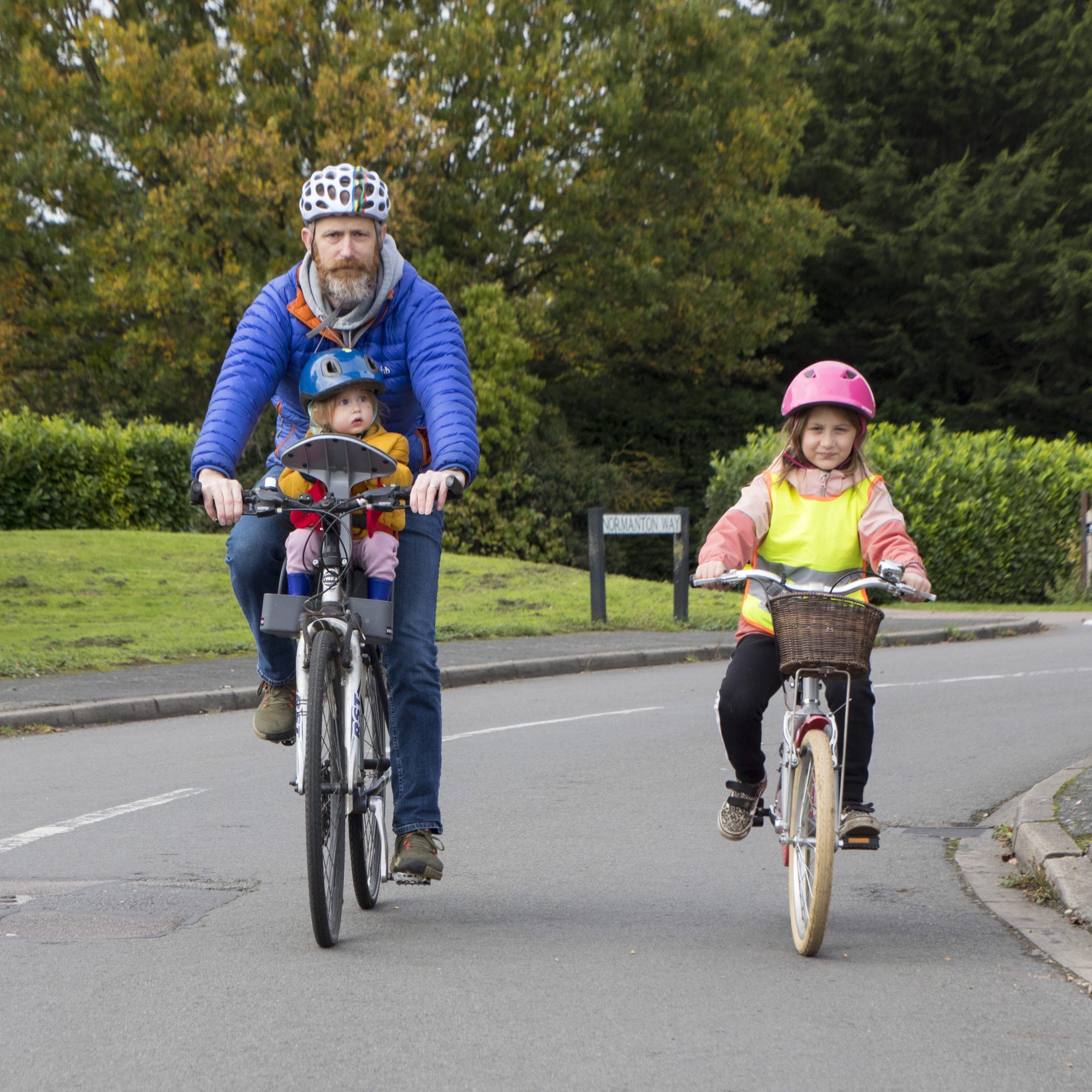A man in a blue jacket and grey helmet rides a bike towards the camera. A toddler is sat in a handlebar seat in front of him. To his right is a young girl in a pink coat and helmet, riding a bike towards the camera.