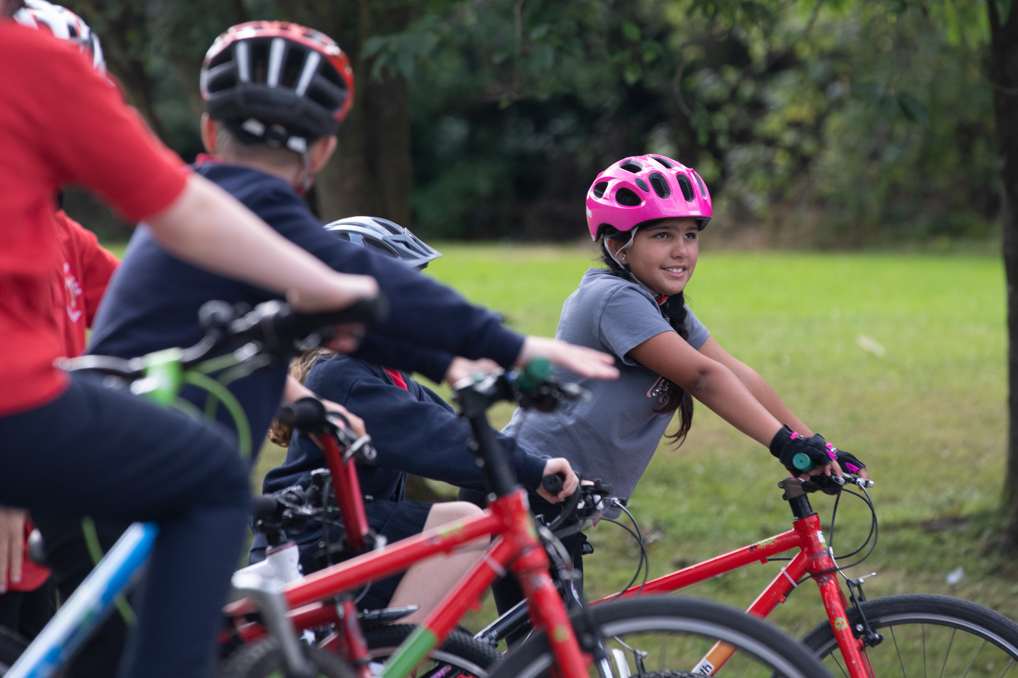 A young girl wearing a pink helment, grey t shirt and standing over a red bike smiles and looks up. In the foreground are other children on bikes looking away from the camera in blur.