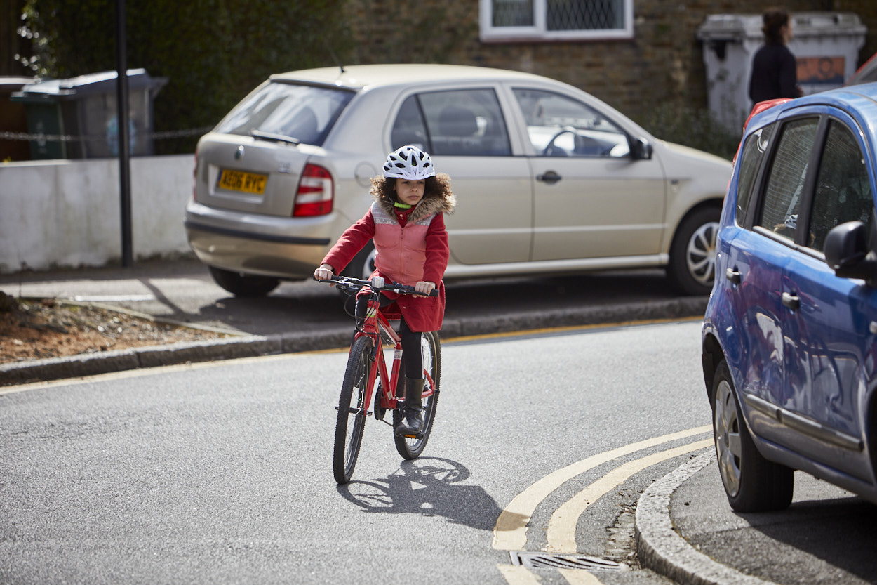 A young girl cycles towards the camera around a bend in the road. She is wearing a red top, pink gilet and a cycling helmet.