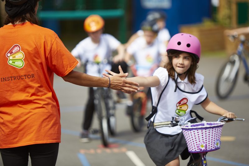 A woman wearing an orange instructor t shirt faces away from the camera and holds her hand out as a young girl wearing a pink helmet and school uniform cycles towards the instructor to high five.