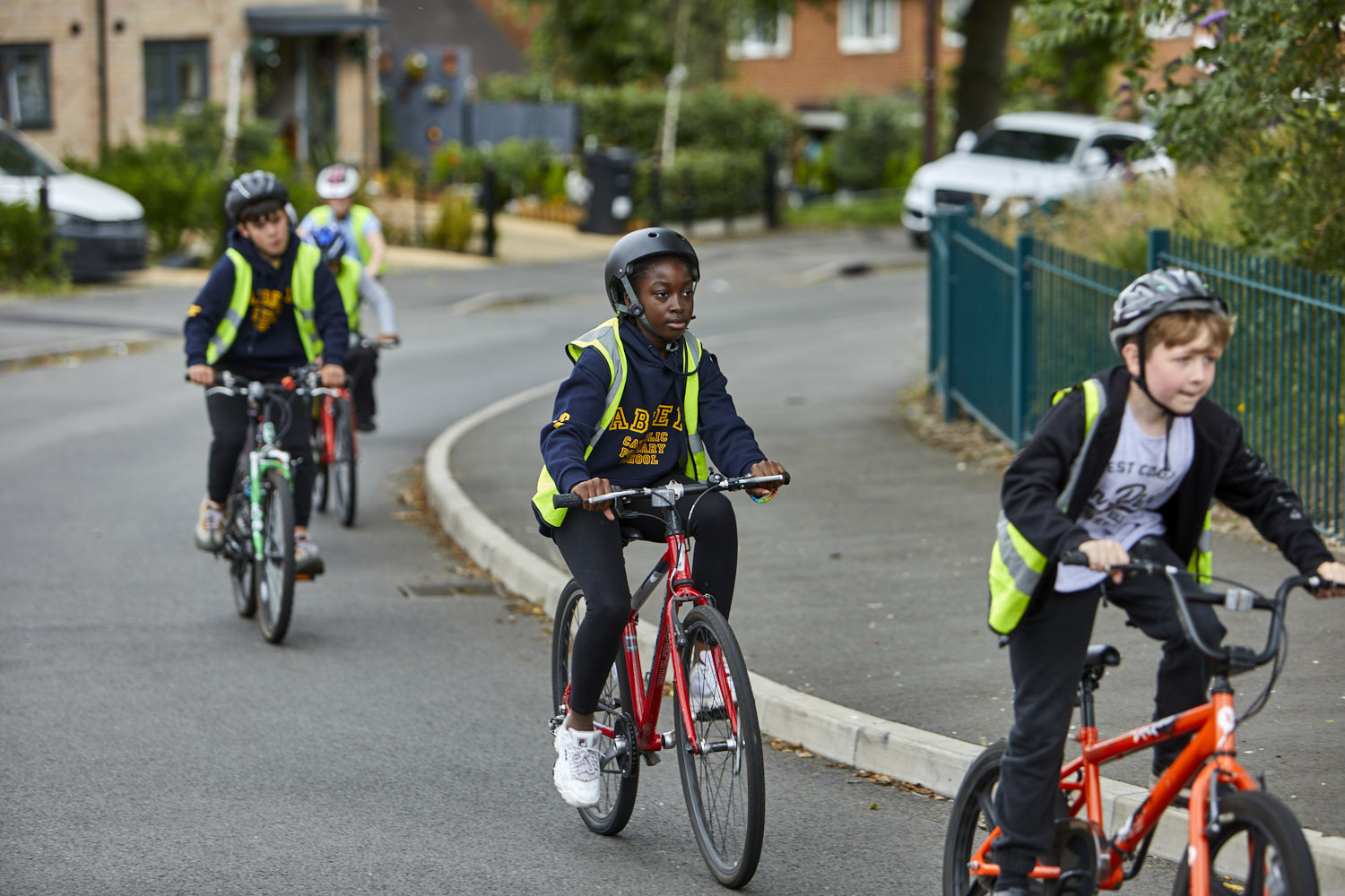 A group of children cycle round the corner of a road, they are wearing high vis vests and cycle helmets and are riding in single file.