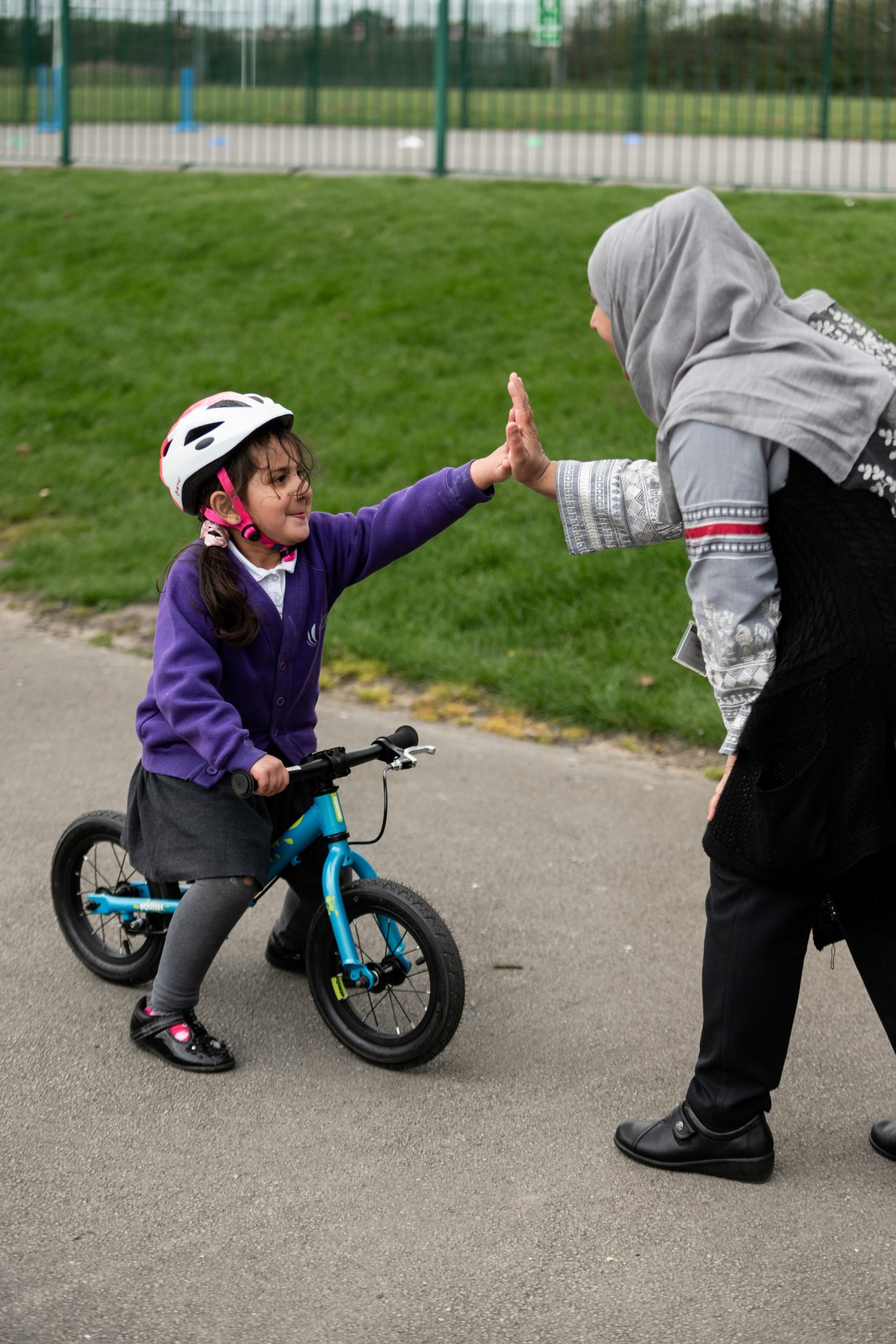 A young girl wearing a purple school cardigan and riding a balance bike high fives a woman wearing a black dress. They are on a playground.