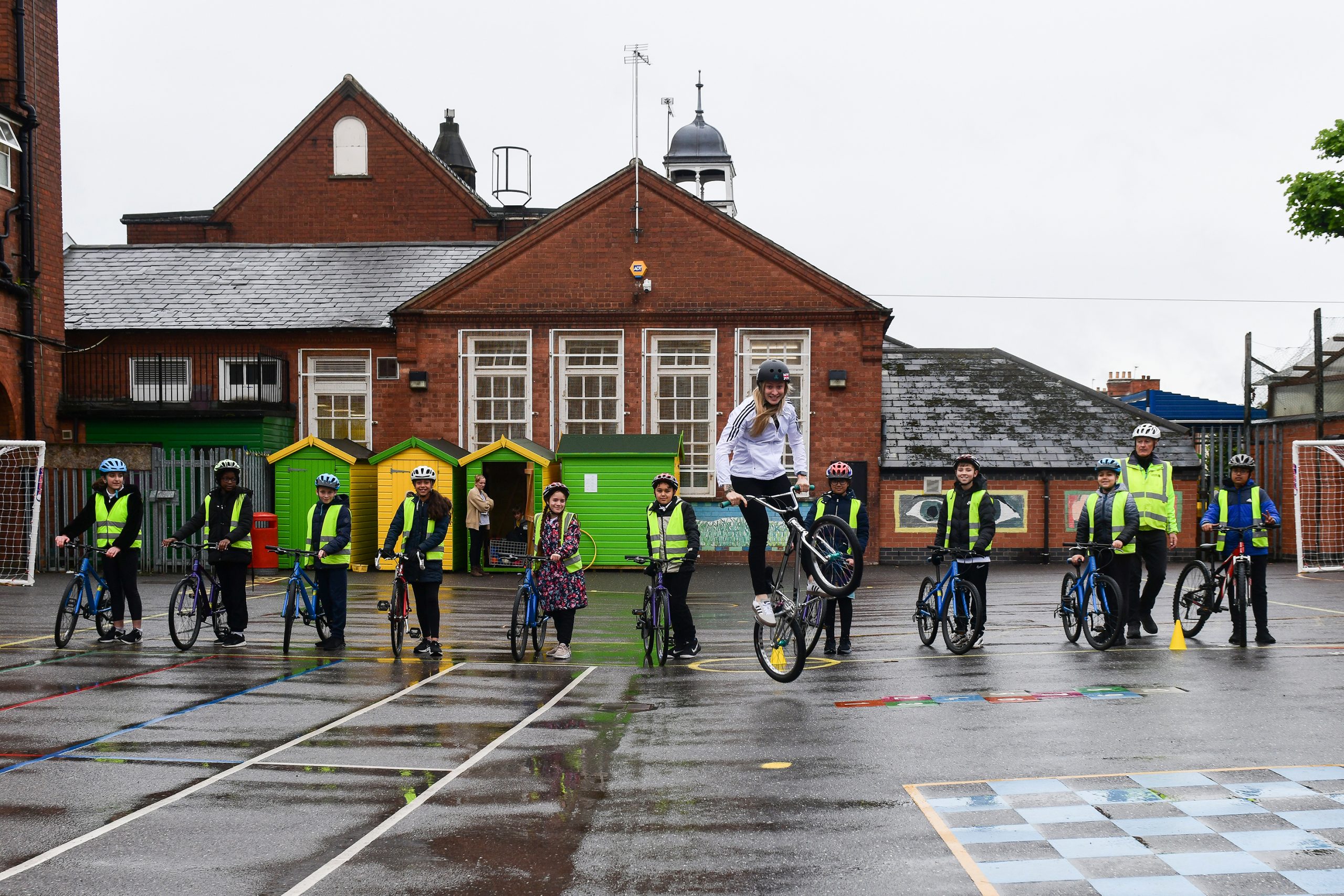 Charlotte Worthingon is performing a BMX stunt in front of a group of school children, standing on a school playground. She is pictured in mid-air, with the group standing behind her smiling in admiration.