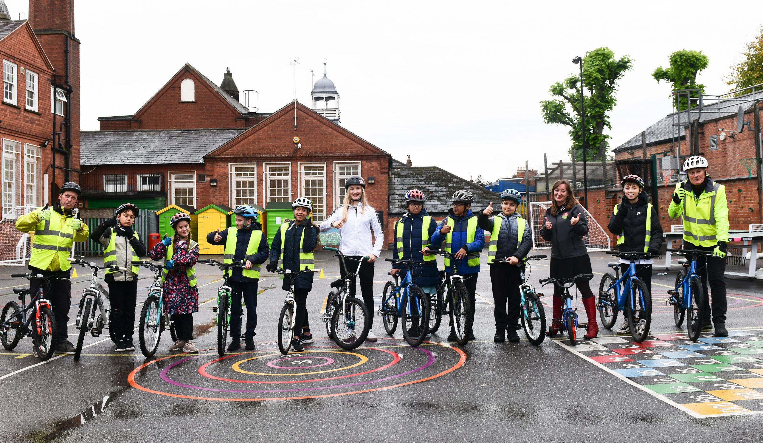 A group of school children are standing in a school playground, with Charlotte Worthington, Emily Cherry, and two Bikeability instructors. They are all wearing safety helmets, and smiling with their thumbs up.