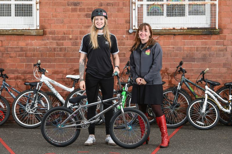 Charlotte Worthington and Emily Cherry are standing with Charlotte's BMX, smiling. A selection of children's bicycles are behind them, against a school wall.