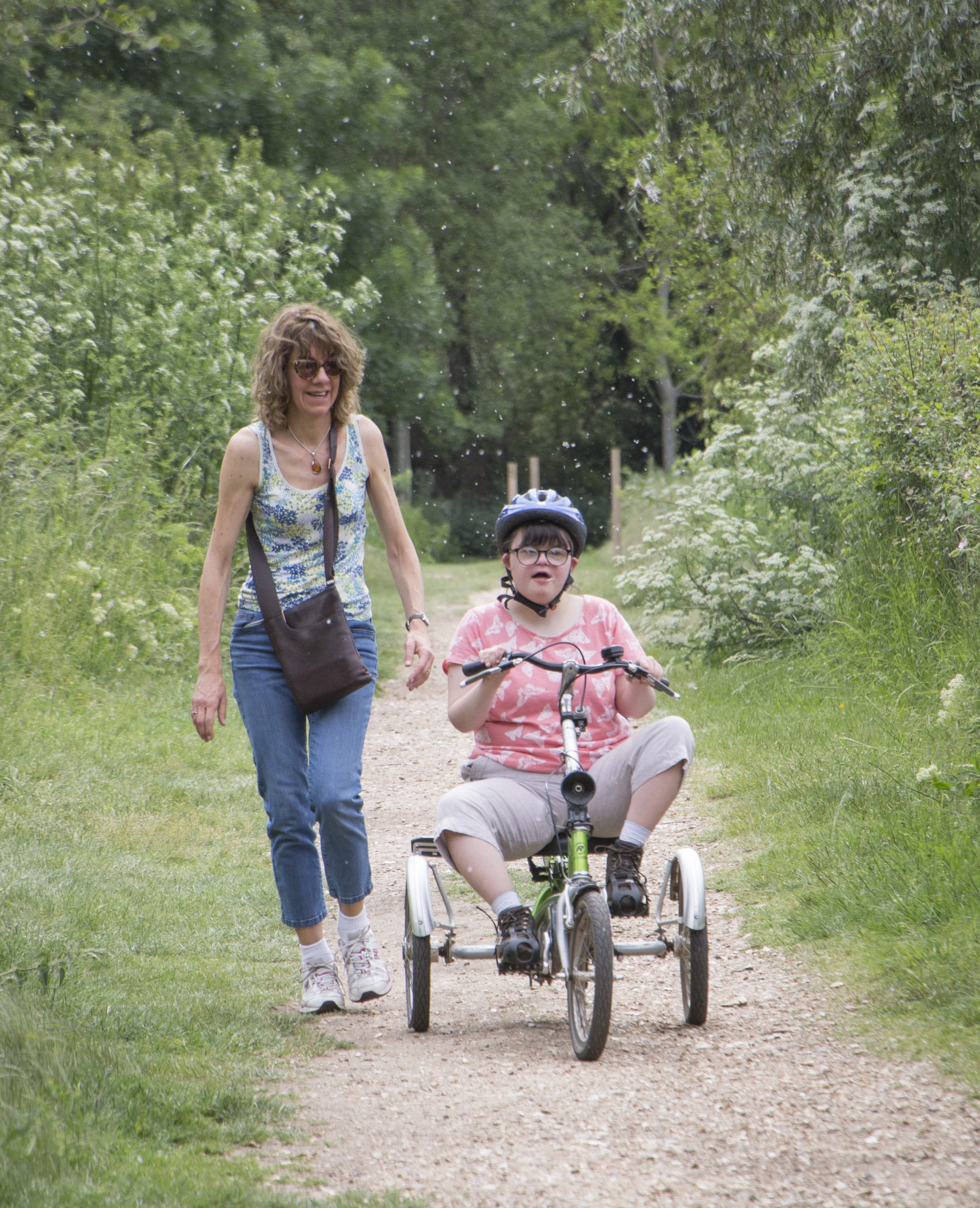 Young cyclist using adapted cycle as her mother walks behind