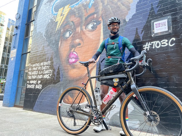 Varun standing with his bike in front of a wall covered in graffiti