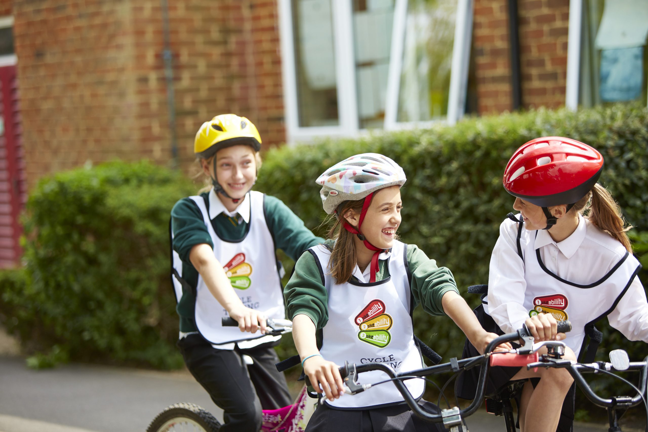 Three girls on cycles and wearing helmets smile at each other. They are stood in front of a building and hedge.