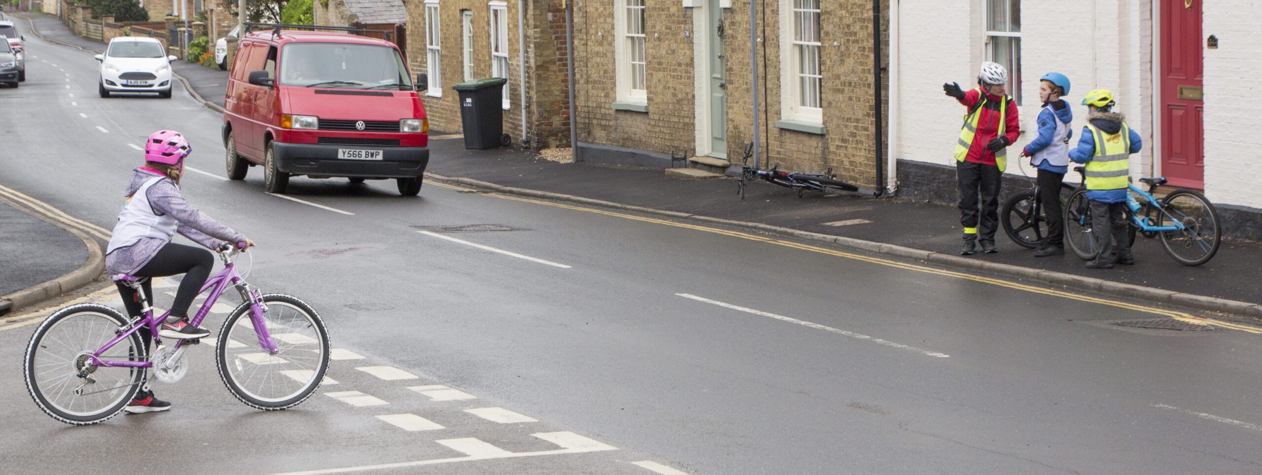 A girl on a pink cycle waits at a t junction, she is wearing a pink helmet. There is a van approaching on the road, driving towards the camera. On the pavement an instructor points out the road layout to two children stood on the pavement.
