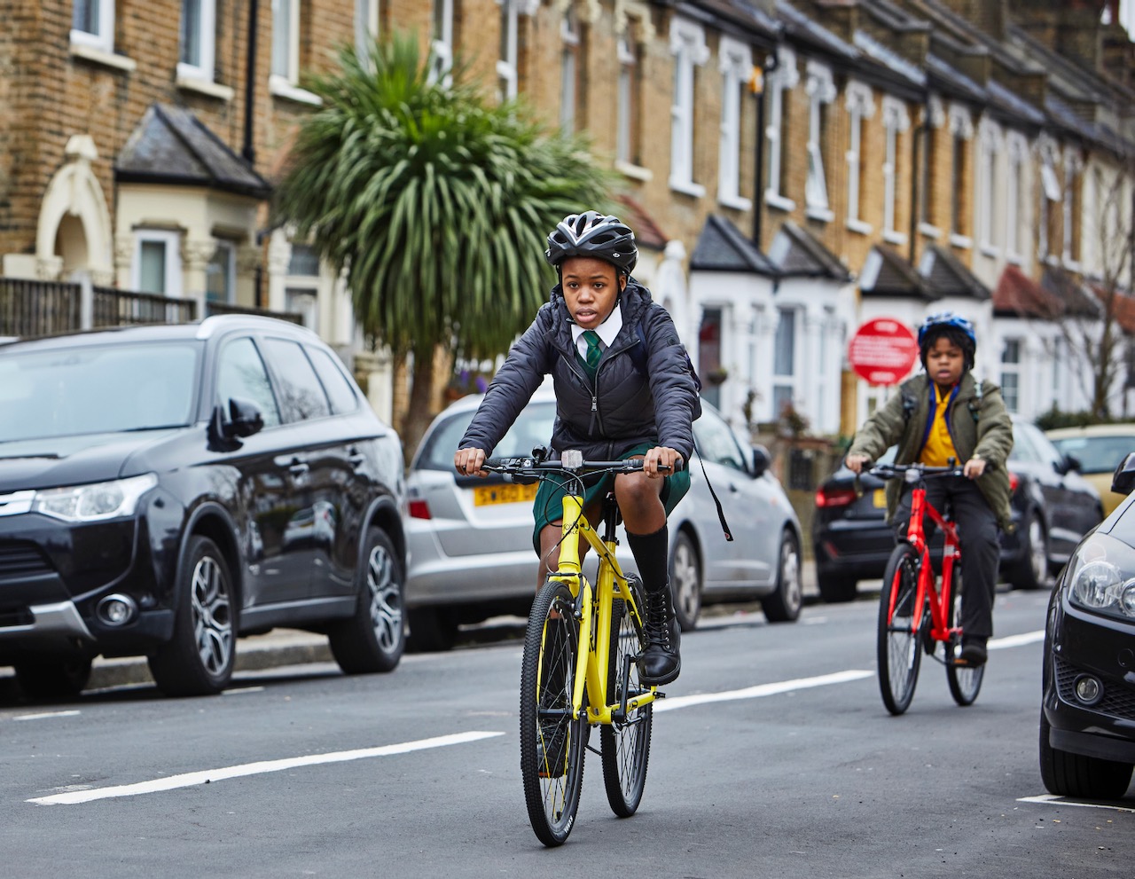 Two children cycling to school on a residential street past parked cars