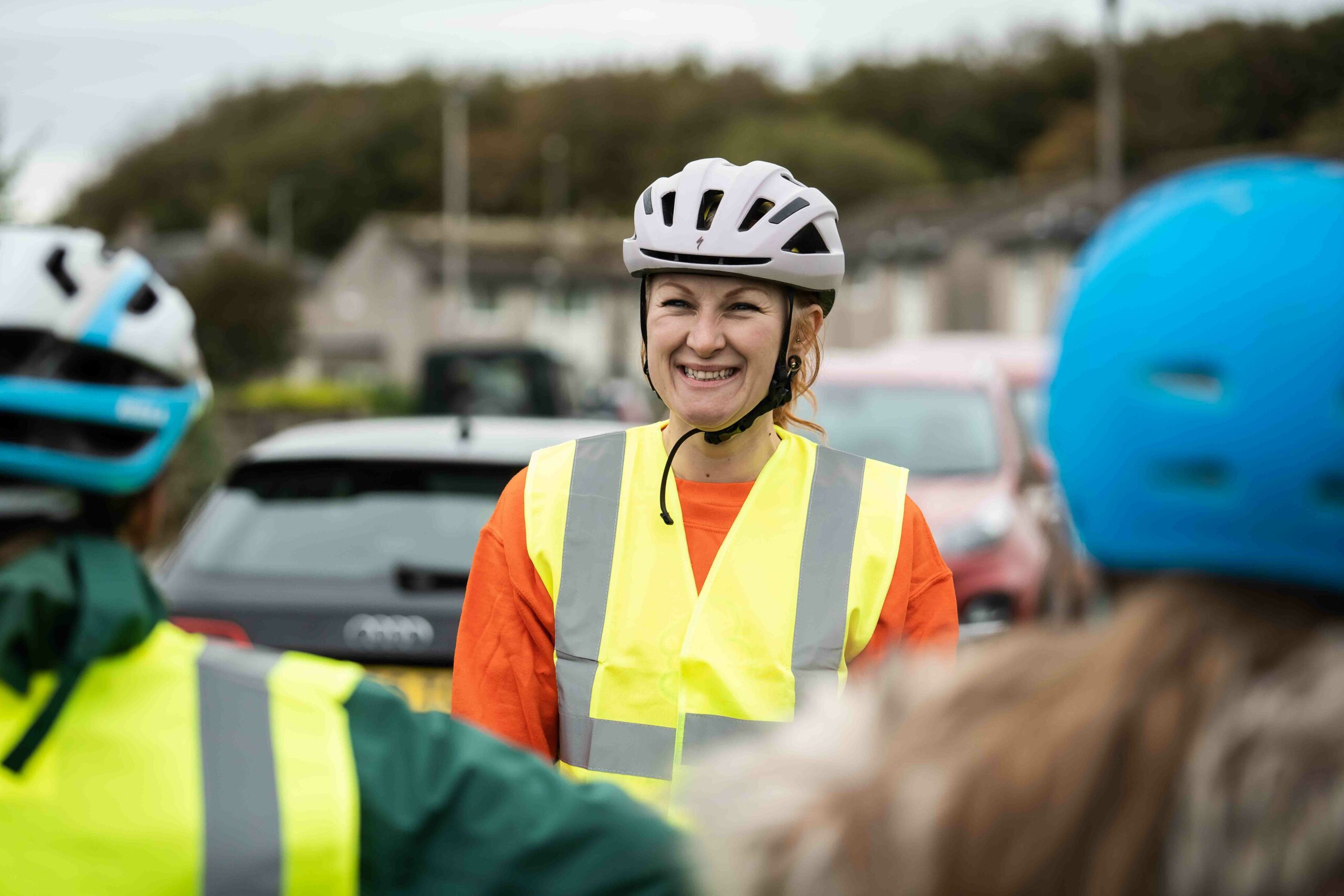 A Bikeability instructor wearing an orange jumper and high vis vest faces the camera smiling. In the foreground are the backs of two children's heads which the camera is looking through their shoulders. .