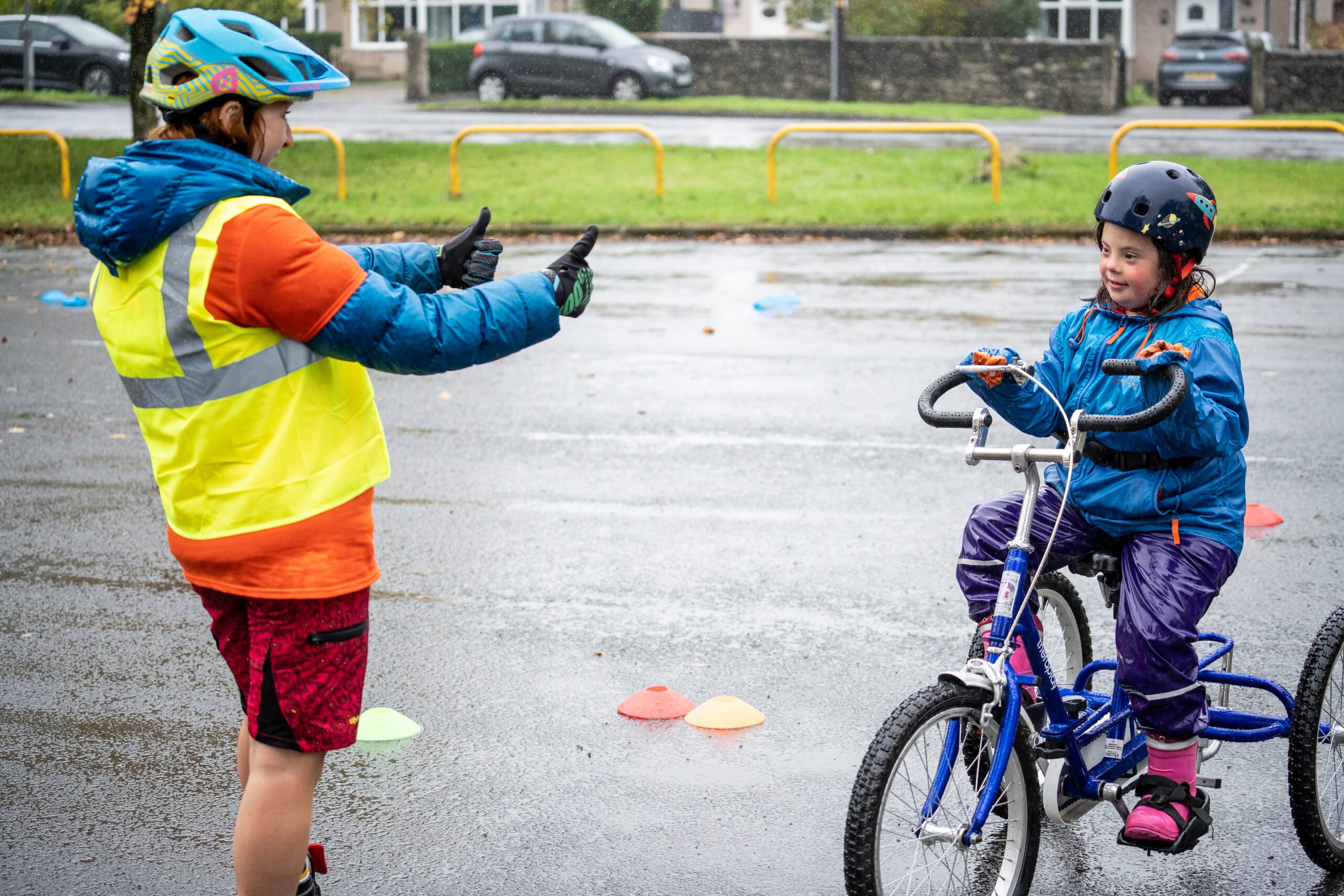 An instructor wearing a blue coat and high vis vest gives the thumbs up to a girl wearing waterproofs and riding a tricycle.