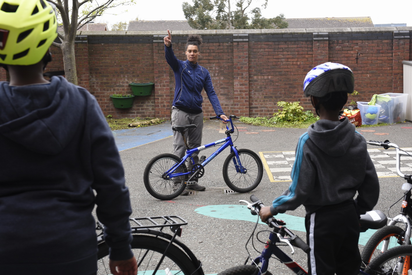 Two children stand by their cycles with their backs to the camera, watching an instructor who is stood by a cycle teaching them.