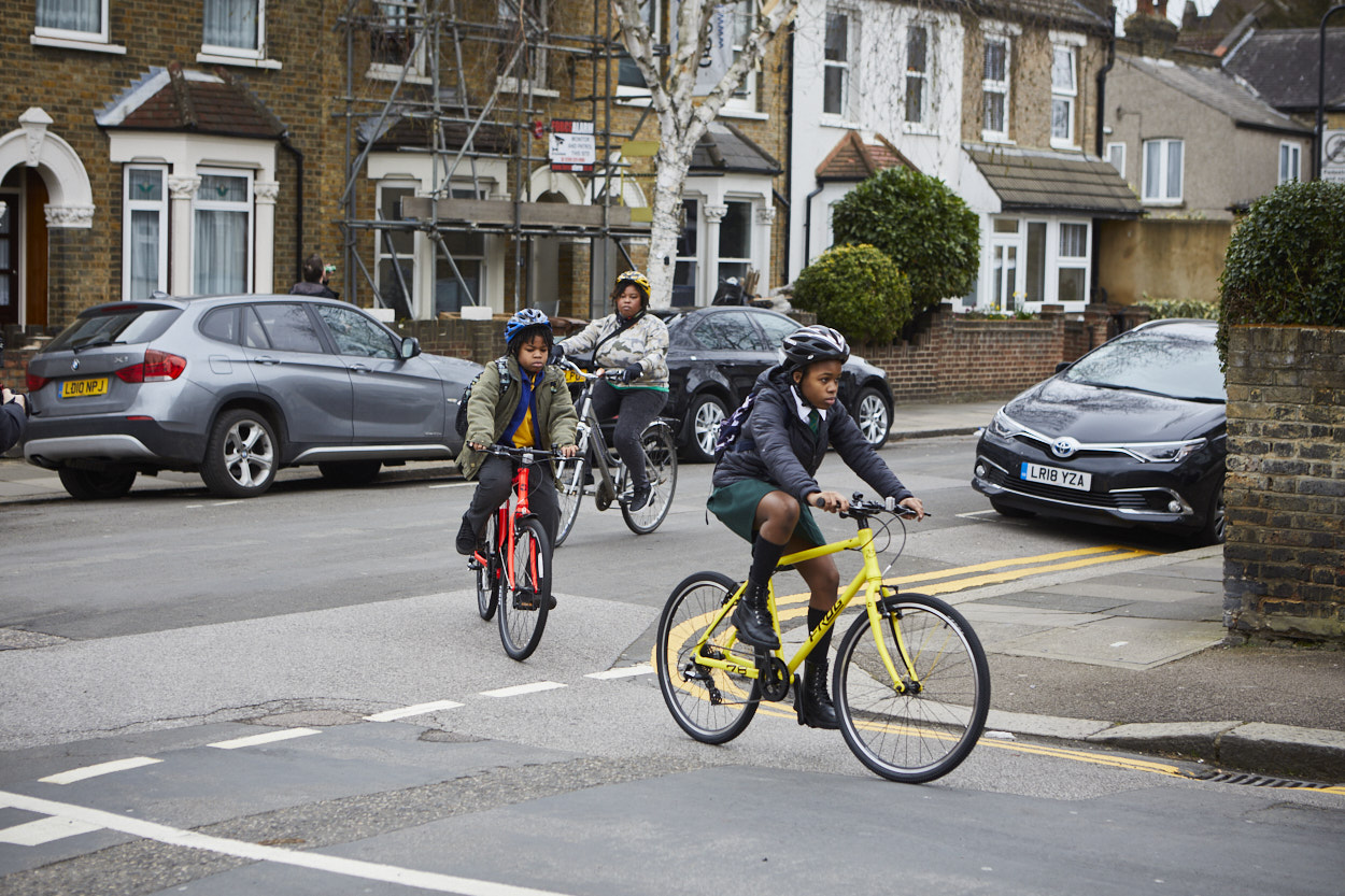 Two children cycle into a side street from a main road. They are wearing coats over their school uniform and are followed by their mother, who is also cycling.