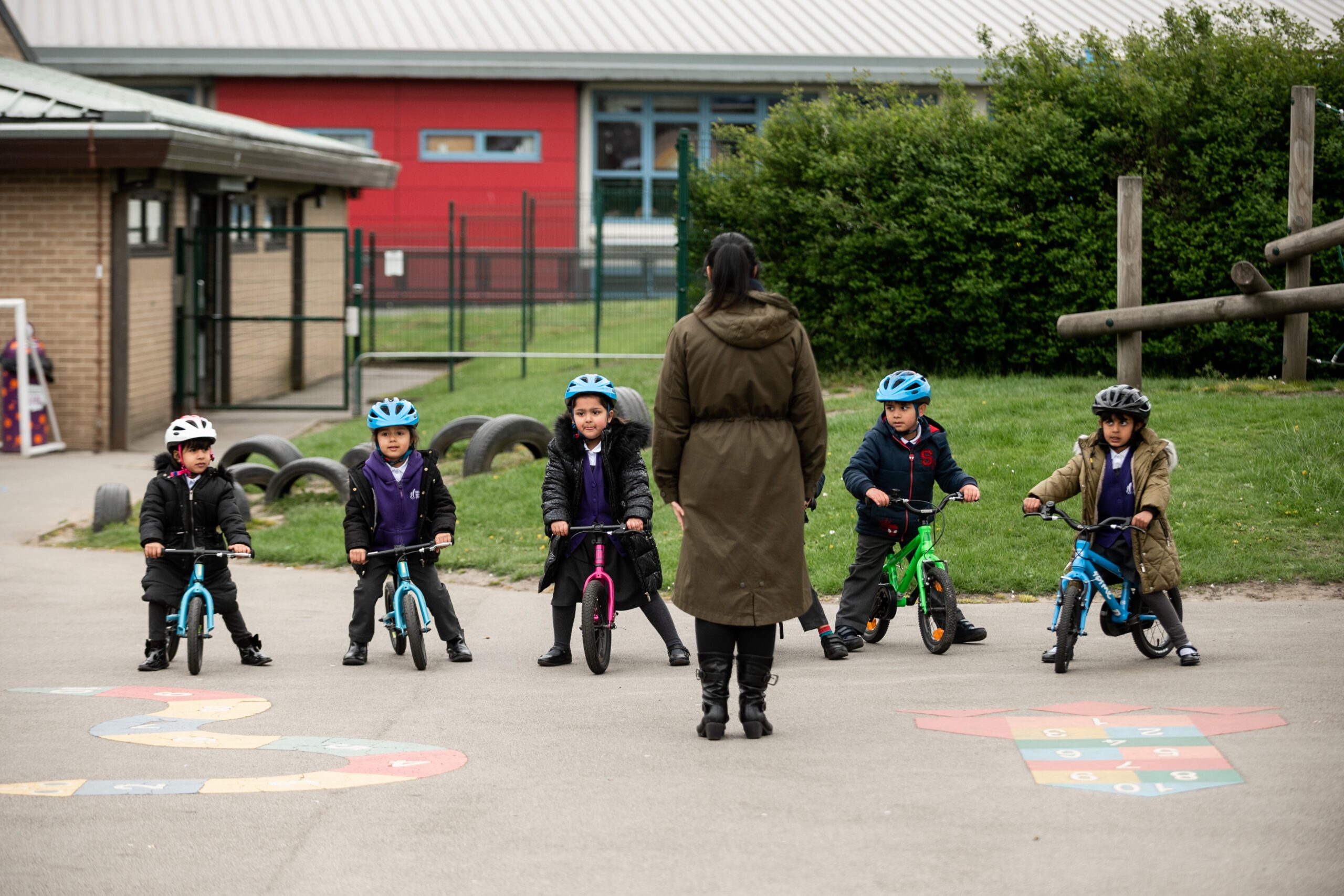 Young school pupils wearing purple school jumpers and sitting on balance bikes are facing the camera, their teacher faces them with her back to the camera. They are on a school playground.