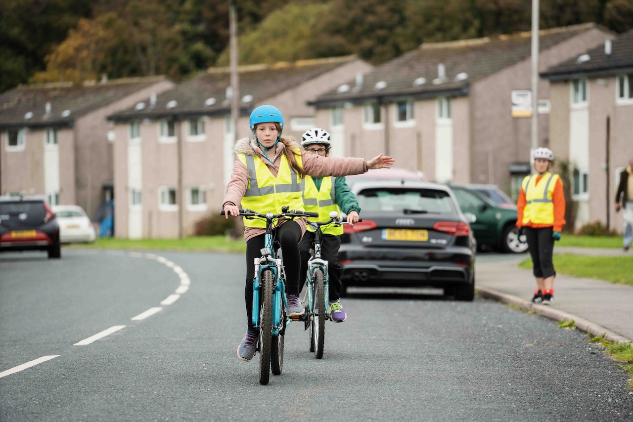 Two Bikeability students practising riding on the road as an instructor watches