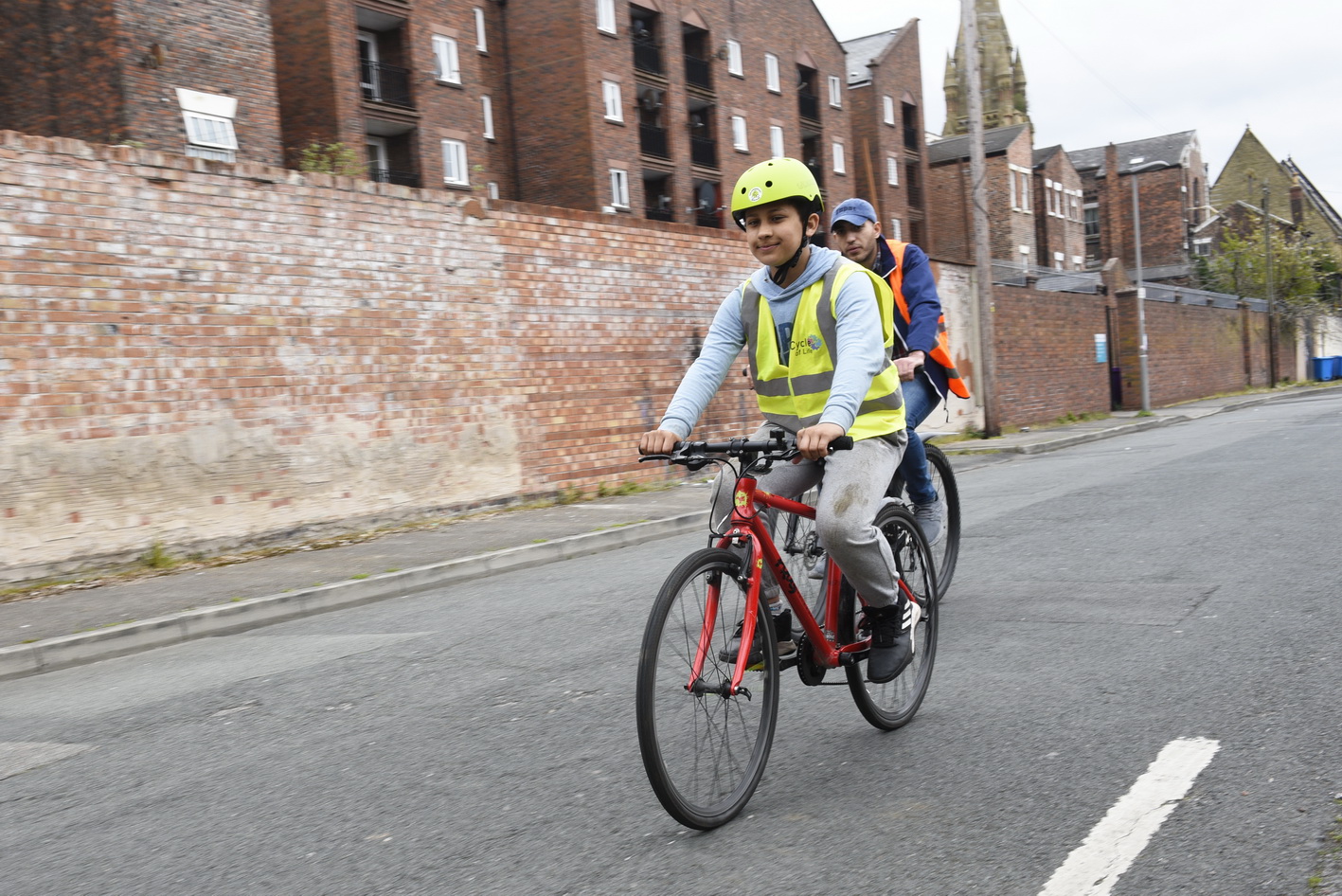 A young boy wearing a high vis vest and riding a red bicycle cycles towards the camera. He is being followed by a man also on a bicycle.. They are cycling on the road.