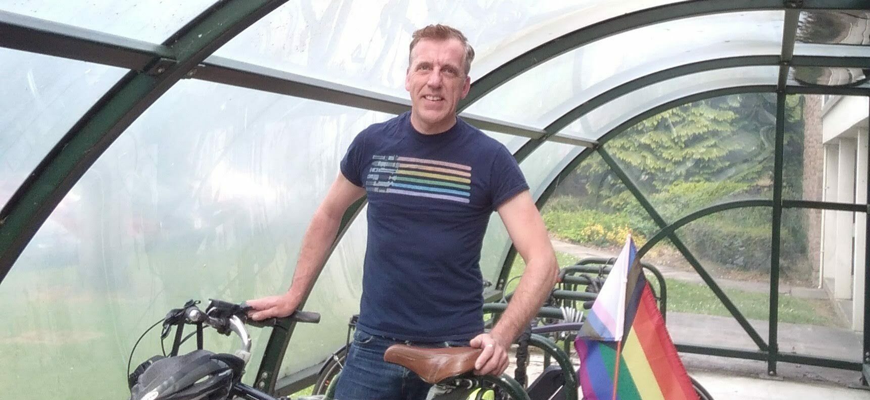 Rob standing behind his bicycle in a bike shelter, a Pride flag in his pannier