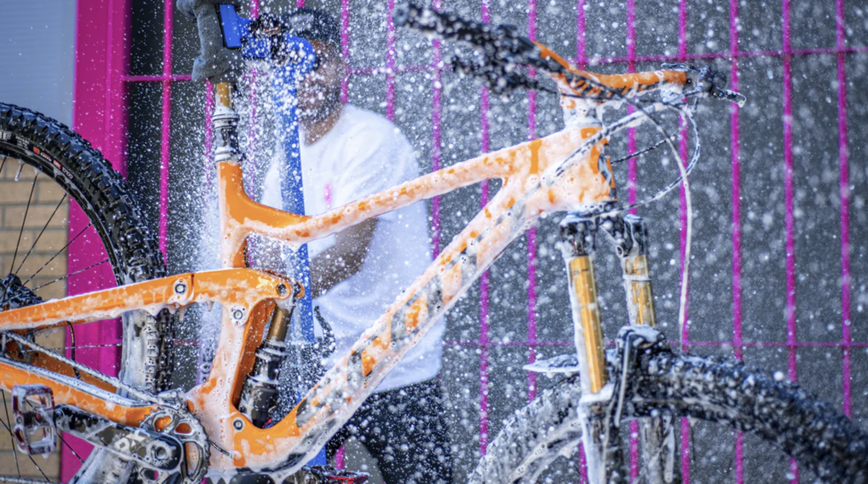 A man standing behind an orange cycle in the forefront of the picture, spraying it with water to clean it