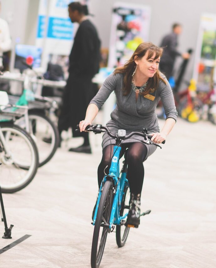 Emily is riding a bicycle inside an exhibition room at the Bikeability Conference.