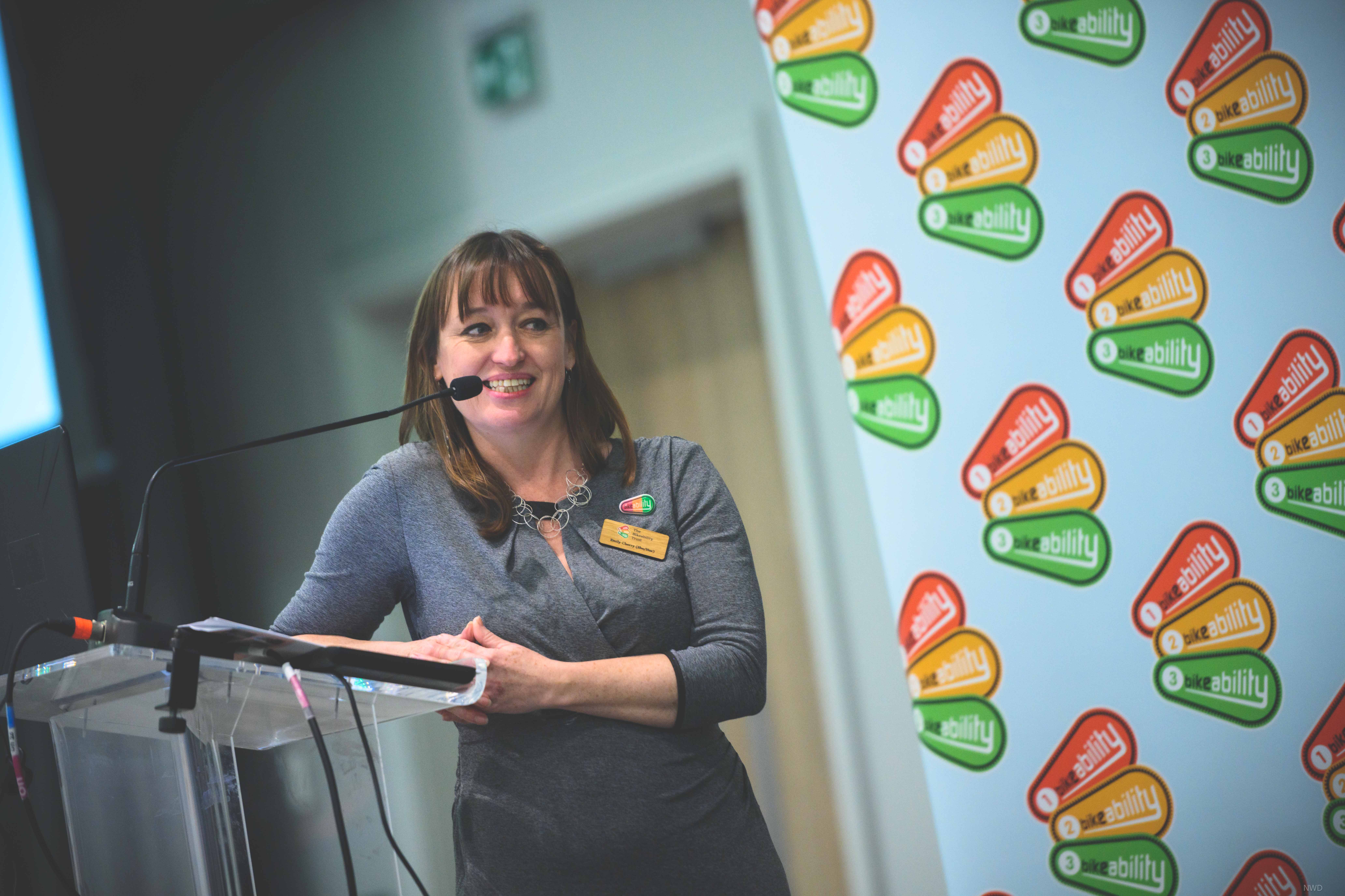 Emily Cherry leans on a lectern, smiling and looking off camera. There is a banner with a repeat pattern of the Bikebaility logo behind her.