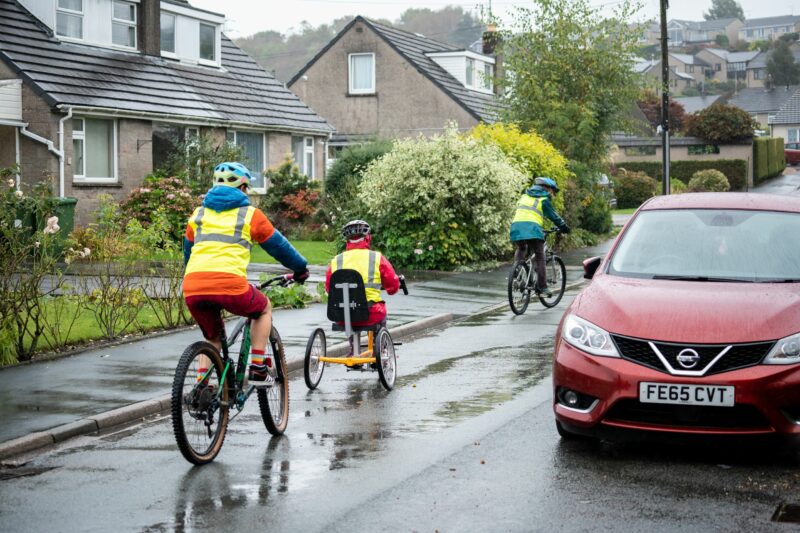 Three people in hi vis cycling away from the camera while on the road. The person in the middle is riding a tricycle