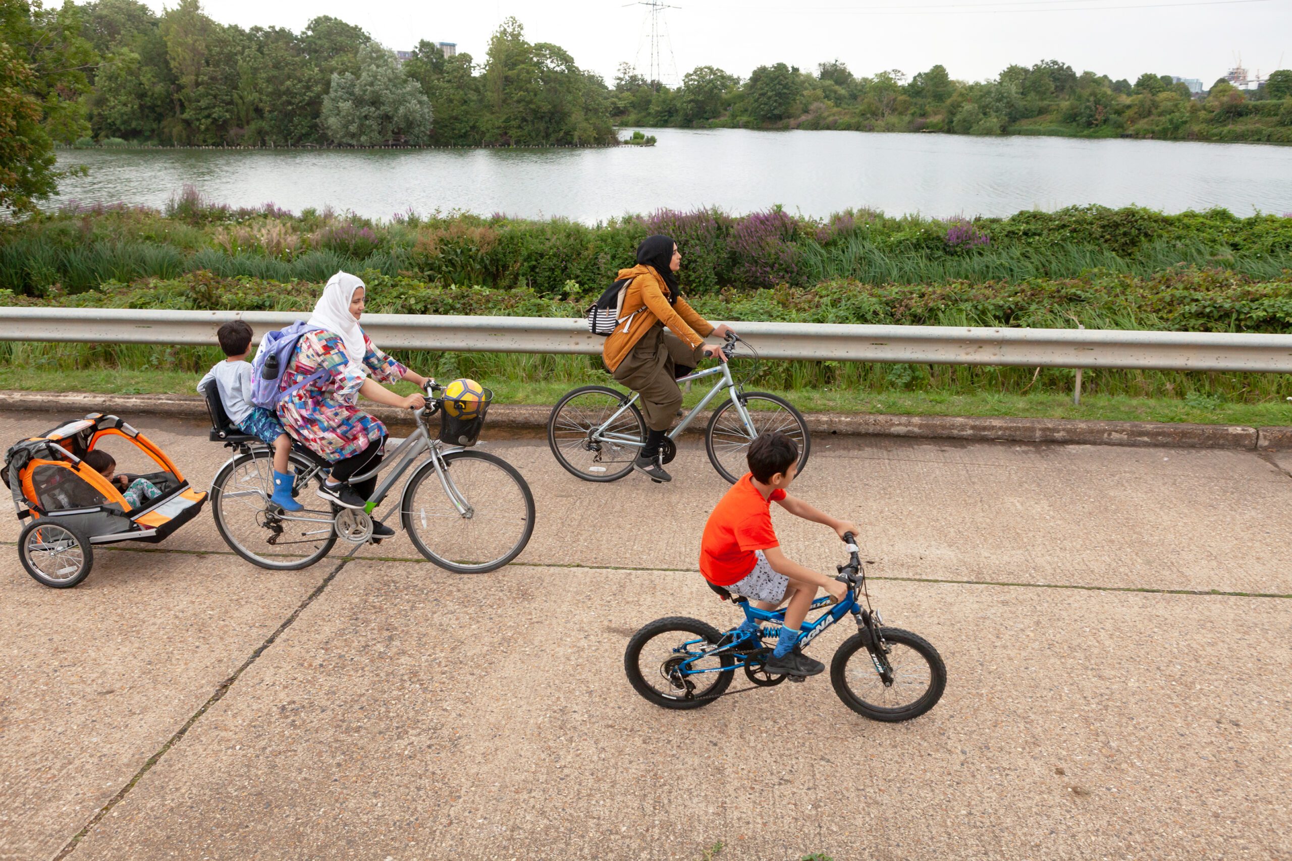 A group of people, including two women in headscarves, a small child on the back of a bike and another in a tag along cycle carrier, plus an older child on his own cycle, cycle past a reservoir