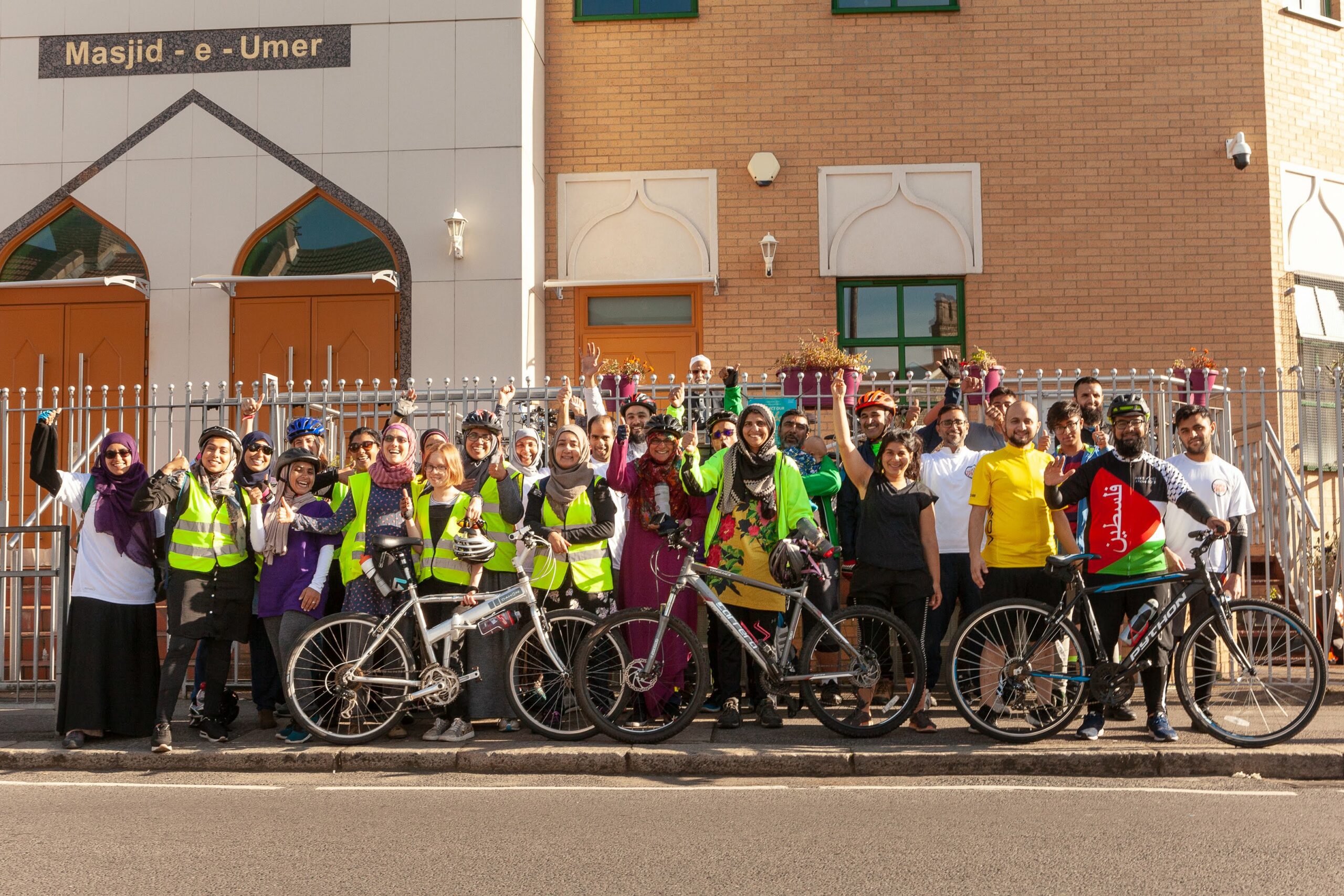 A group of people with bicycles standing outside a Mosque waving