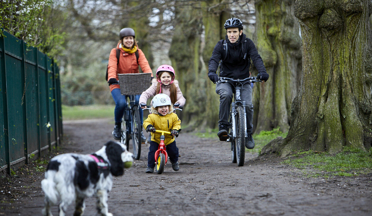 A family of four riding their cycles in the park. There is a dog in the foreground and the family are laughing as they cycle.