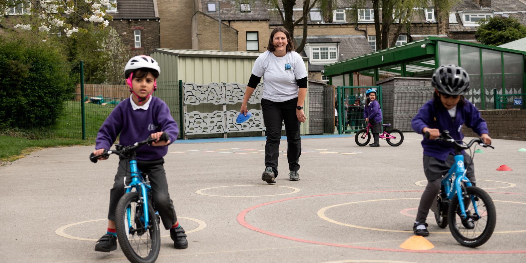 A Bikeability instructor is teaching two young children on balance bikes in a playground