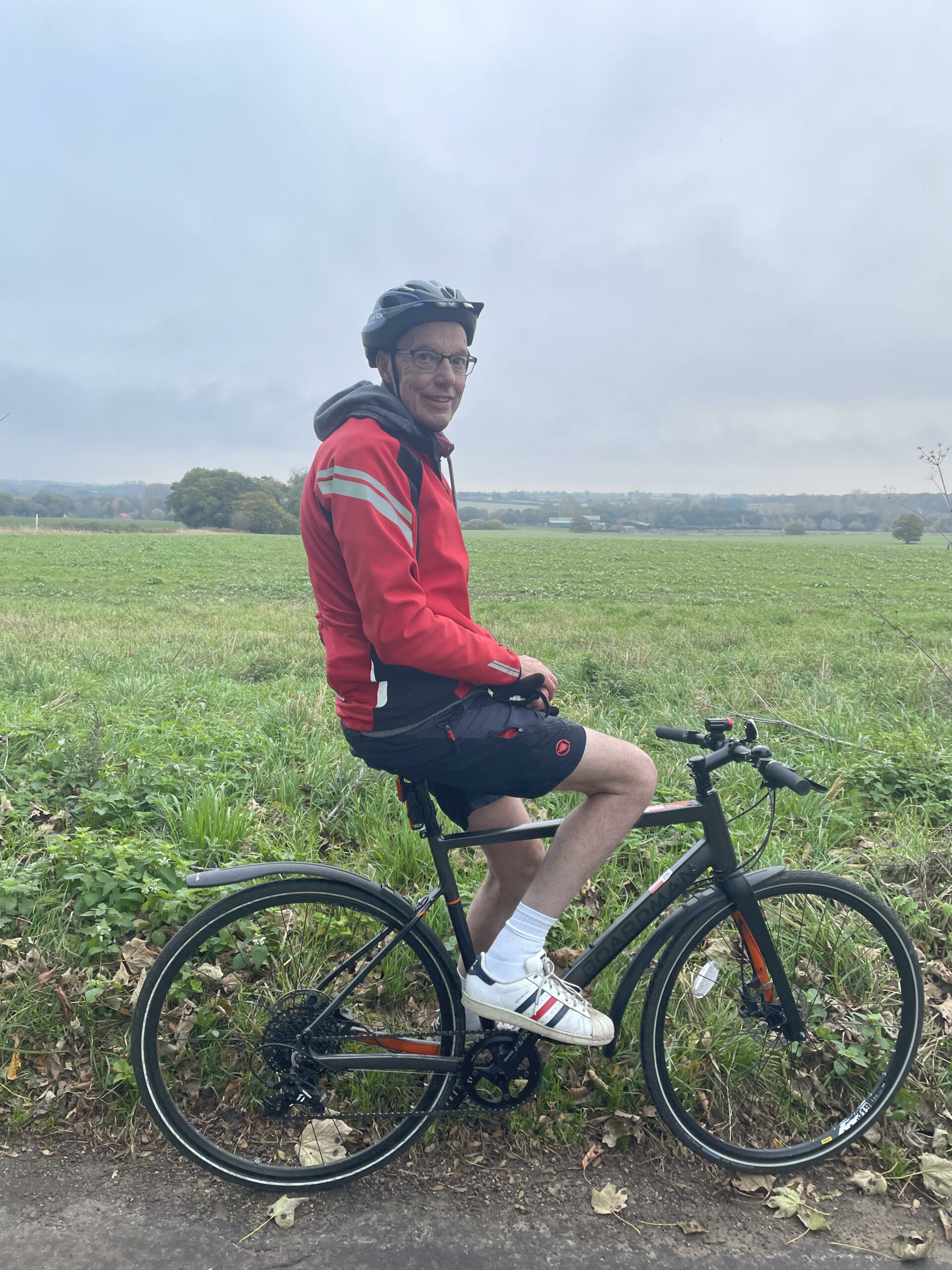 Andrew is sitting on his bicycle by a picturesque field. He is wearing a helmet and smiling.