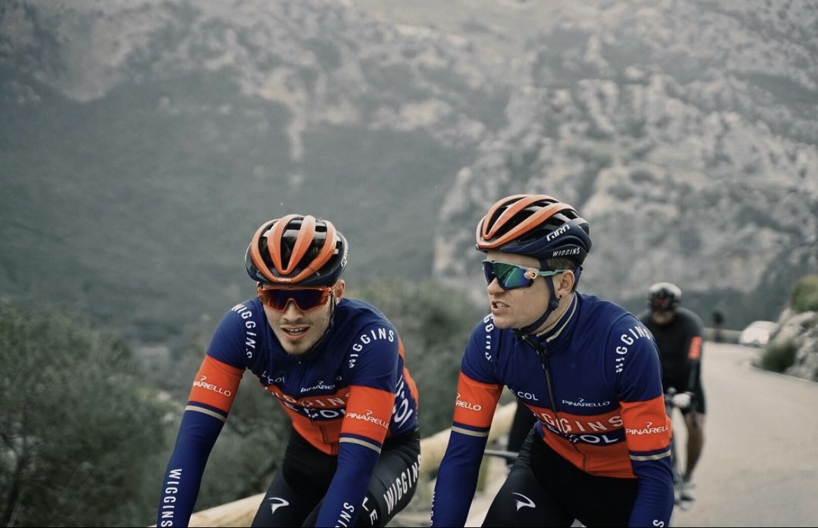 Jacques and a team mate cycling in a road race. There is a mountain in the background and they are wearing blue and red Team Wiggins jerseys and helmets