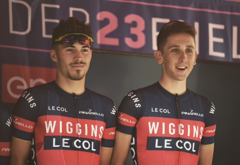 Jacques and team mate standing in front of a race advert back drop. They are both wearing red and blue Team Wiggins jerseys