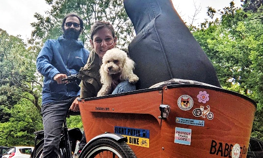 A cargo trike ridden by a white man has a white woman with a feeding tube, a poodle and a cello in the cargo box on the front