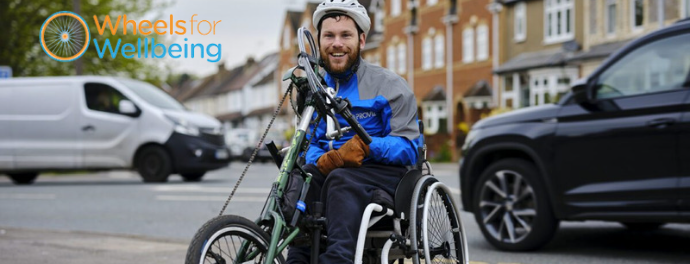 A man is sitting on an adapted hand cycle. He is smiling and wearing a safety helmet. The Wheels for Wellbeing logo is in the top left corner.